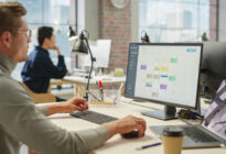 Junger Mann sitzt im Büro am Laptop und am Computer an einer Projektmanagementsoftware