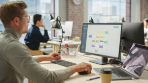 Junger Mann sitzt im Büro am Laptop und am Computer an einer Projektmanagementsoftware